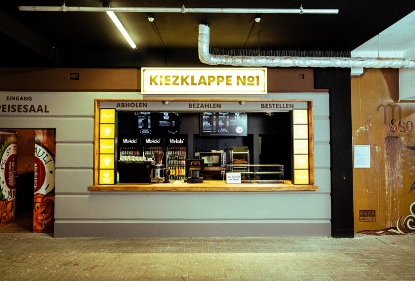 Ein Kiosk mit dem Namen 'Kiezklappe No1' im Stadion oder Vereinsgelände des FC St. Pauli. Der Kiosk hat ein Schild mit den Bereichen 'Abholen', 'Bezahlen' und 'Bestellen'. Im Hintergrund sind verschiedene Getränke und Snacks zu sehen, darunter auch Fitz-Kola. Links neben dem Kiosk befindet sich ein Eingang mit einem Schild 'Eingang Speisesaal' und einem dekorativen Element mit dem Logo des FC St. Pauli.