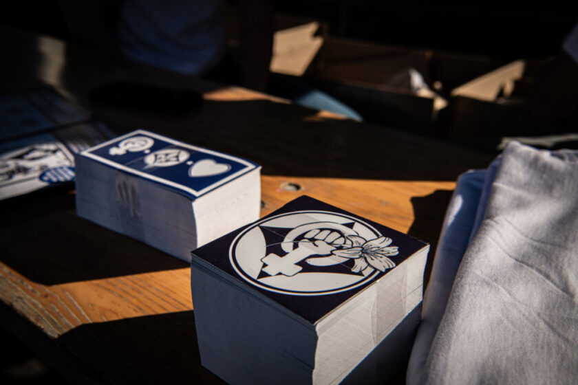 Drei Holzkisten mit weißem und blauem Design, die auf einem Holztisch stehen. Die Kisten sind mit einem Logo verziert, das eine Hand mit einem Blatt darin zeigt. Im Hintergrund sind unscharfe Gegenstände und eine Person zu erkennen.