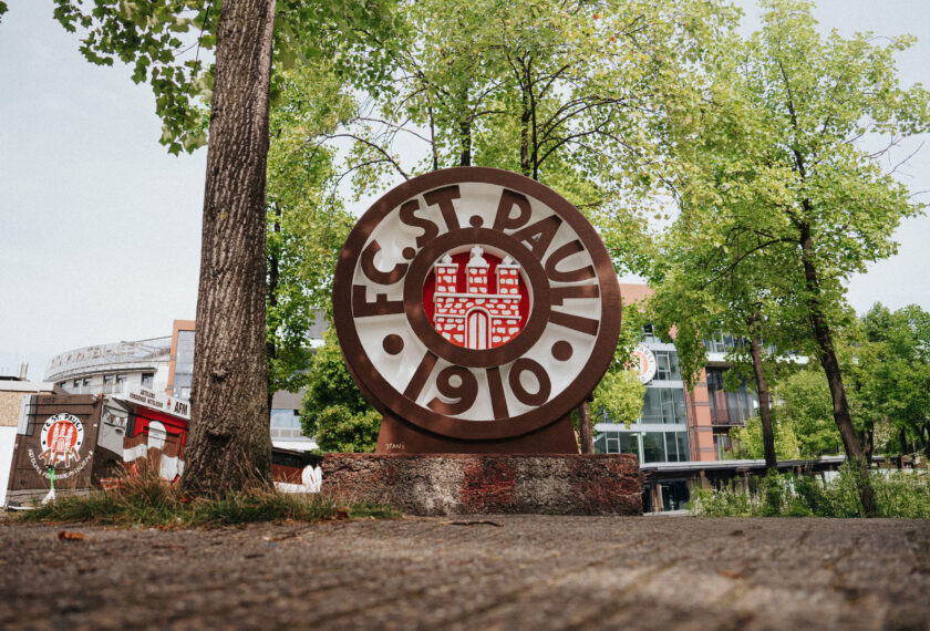 Ein großes, rundes Steinwappen des FC St. Pauli mit dem Vereinslogo und der Jahreszahl 1910, umgeben von Bäumen und Gebäuden im Hintergrund.