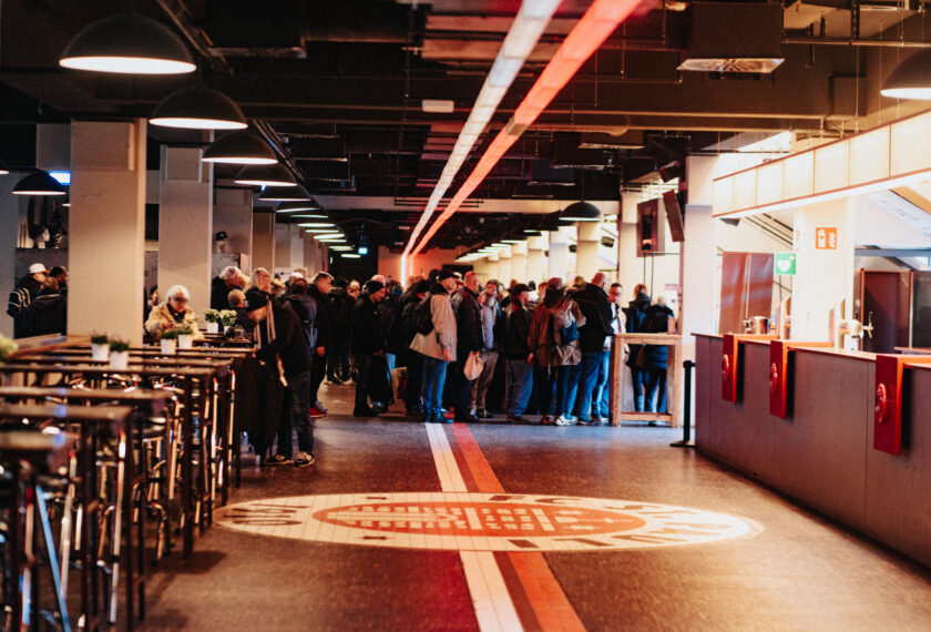 Menschen stehen in einer Schlange in einem gut beleuchteten Innenraum, möglicherweise in einem Stadion oder einer Arena. Der Boden hat ein markantes Design mit dem Logo des FC St. Pauli. Die Atmosphäre wirkt lebhaft und gesellig, was auf eine Veranstaltung oder ein Treffen hinweist.