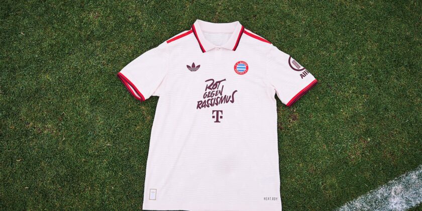 Ein weißes Trikot des FC Bayern München liegt auf einem Rasen. Das Trikot hat rote und schwarze Akzente und zeigt das Logo des FC Bayern München, den Adidas-Logo und den Slogan "Rot gegen Rassismus".