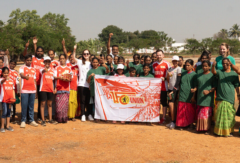 Eine Gruppe von Menschen, darunter Kinder und Erwachsene, steht auf einem Sportplatz und hält ein Banner mit dem Logo von Hertha BSC und dem Text "Hertha BSC Union". Die Personen tragen verschiedene T-Shirts, einige mit den Aufschriften "Berlin" und "EFLIN". Sie posieren für ein Gruppenfoto und wirken fröhlich und engagiert.