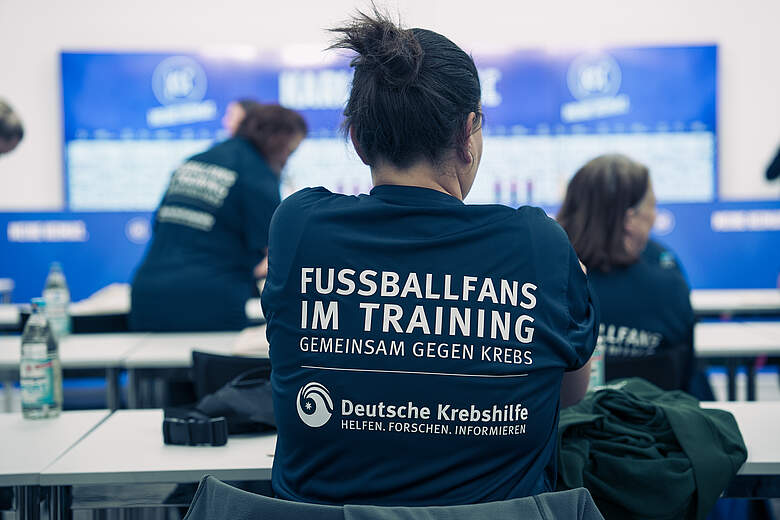 Eine Person mit einem T-Shirt, das den Text "FUSSBALLFANS IM TRAINING GEMEINSAM GEGEN KREBS" und das Logo der Deutschen Krebshilfe trägt, sitzt an einem Tisch in einem Raum mit mehreren anderen Personen. Im Hintergrund sind Bildschirme mit Informationen zu sehen.