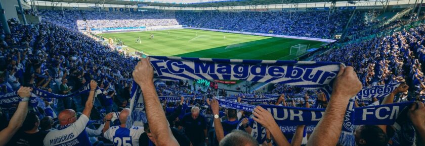 Ein Panoramafoto eines voll besetzten Fußballstadions. Fans im Vordergrund halten ein großes blau-weißes Transparent mit der Aufschrift "FC Magdeburg". Die Spieler sind auf dem grünen Rasen zu sehen, und die Atmosphäre ist lebhaft und energiegeladen.
