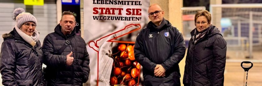 Vier Personen stehen vor einem Plakat, das gegen Lebensmittelverschwendung aufruft. Das Plakat zeigt einen Einkaufswagen mit Obst und den Text "Lebensmittel statt sie wegzuwerfen!". Die Personen sind warm gekleidet und lächeln in die Kamera.