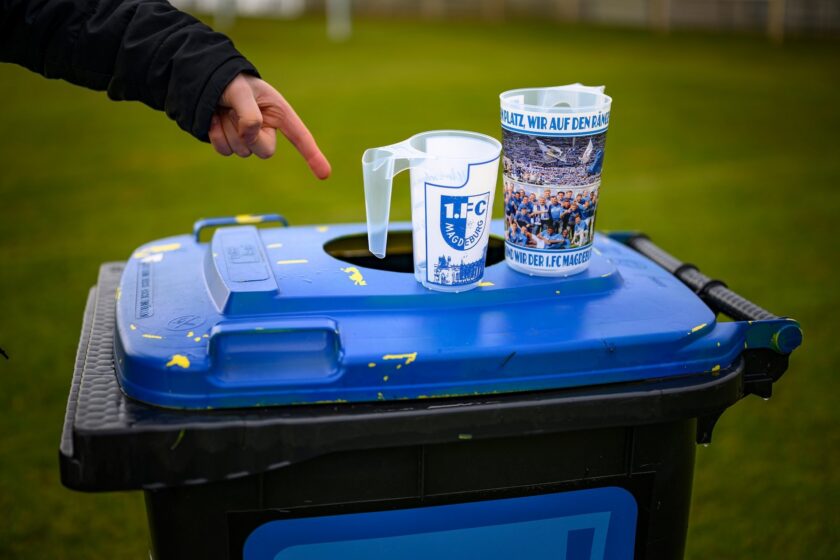Eine Hand zeigt auf zwei Plastikbecher, die auf einem blauen Müllcontainer liegen. Die Becher tragen Aufschriften und Logos des 1. FC Magdeburg. Im Hintergrund ist ein Rasenplatz zu sehen.