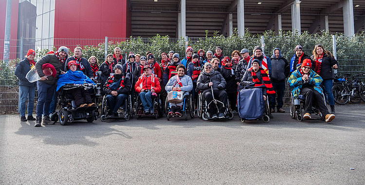 Eine Gruppe von Menschen, darunter einige im Rollstuhl, stehen vor einem Stadion und lächeln in die Kamera. Sie tragen verschiedene farbige Kleidung, darunter einige mit Schal und Mütze. Die Atmosphäre wirkt fröhlich und gesellig.