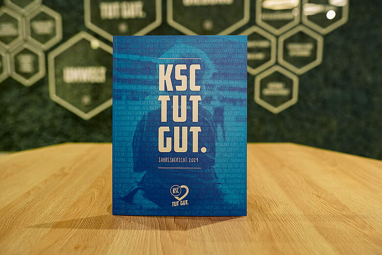 Ein blaues Cover eines Jahresberichts des KSC (Karlsruher SC) mit dem Slogan "TUT GUT." und dem Jahr 2023. Im Hintergrund sind hexagonale Elemente mit dem Vereinslogo zu sehen.