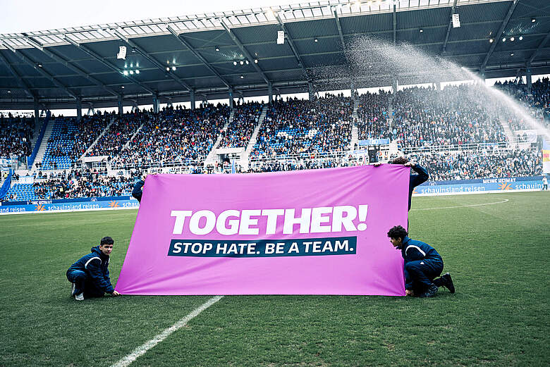 Zwei Personen halten ein großes pinkes Banner auf einem Fußballfeld, auf dem "TOGETHER! STOP HATE. BE A TEAM." steht. Im Hintergrund sind viele Zuschauer auf den Tribünen zu sehen.