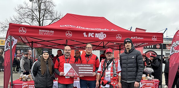 Eine Gruppe von Menschen steht vor einem roten Zelt mit dem Logo des 1. FC Nürnberg und dem Slogan "Bundesliga bewegt". Sie halten einen Scheck über 800 Euro in den Händen, der für das Projekt "Gemeinsam gegen Armut" bestimmt ist. Die Personen tragen rote Kleidung und lächeln in die Kamera. Im Hintergrund sind weitere Zelte und Menschen zu sehen.
