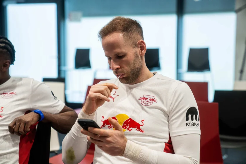 Zwei Männer in weißen T-Shirts mit dem Logo des RB Leipzig sitzen in einem Raum und nutzen ihre Smartphones. Einer der Männer trägt ein blaues Armband und hat eine Verletzung am Arm, die bandagiert ist. Im Hintergrund sind Fenster mit Jalousien zu sehen.