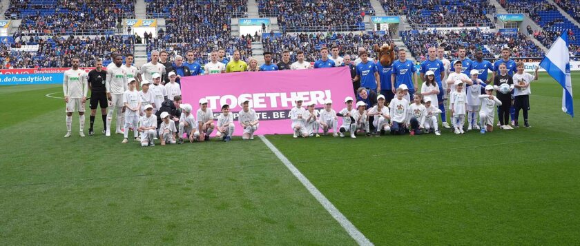 Gruppenfoto von Spielern, Kindern und Vereinsmaskottchen auf einem Fußballfeld. Sie halten ein großes Transparent mit der Aufschrift 'TOGETHER! RESPECT. UNITE. BE A HERO.' Die Spieler und Kinder tragen weiße und blaue Trikots. Im Hintergrund sind viele Zuschauer auf den Tribünen zu sehen.