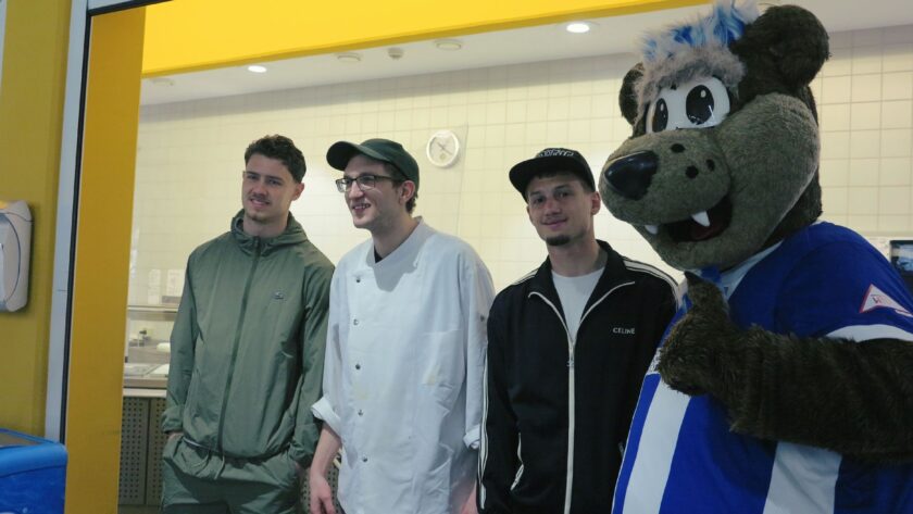 Drei Männer und ein Maskottchen stehen in einem Raum, der durch eine Glaswand getrennt ist. Die Männer lächeln und scheinen sich zu unterhalten. Einer der Männer trägt eine grüne Jacke, der andere eine weiße Schürze und der dritte eine schwarze Jacke mit einem schwarzen Cap. Das Maskottchen, das wie ein Bär aussieht, trägt eine blaue Jacke mit weißen Streifen und einem Logo auf der Brust. Im Hintergrund sind medizinische Geräte und eine Uhr zu sehen.