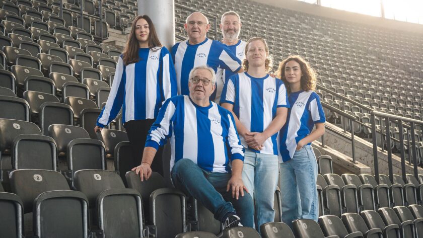 Eine Gruppe von fünf Personen sitzt auf den Tribünen des Olympiastadions und trägt blaue und weiße Trikots des Hertha BSC. Im Hintergrund sind leere Sitzreihen zu sehen.