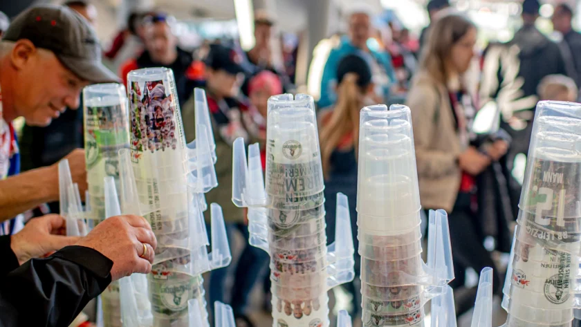 Menschen in einer Menschenmenge, die durchsichtige Plastikbecher mit bedruckten Designs halten. Die Becher sind stapelbar und haben verschiedene Aufdrucke, darunter Logos und Bilder. Die Szene scheint bei einem öffentlichen Event oder einer Veranstaltung zu sein.