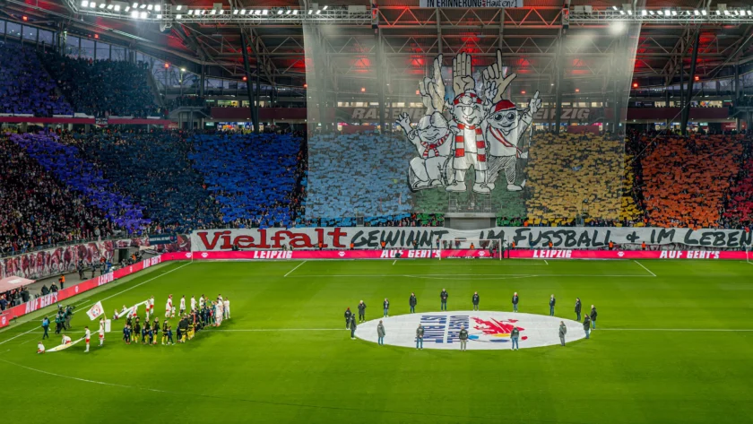 Ein Stadion voller Fans, die ein Fußballspiel zwischen RB Leipzig und Borussia Dortmund erwarten. Im Hintergrund ist eine große Choreografie mit drei Figuren zu sehen, die die Hände in die Luft strecken. Vor den Fans steht ein Transparent mit der Aufschrift "Vielfalt gewinnt im Fußball und im Leben". Auf dem Spielfeld sind die Mannschaften und Schiedsrichter in der Mitte versammelt, bereit für den Anpfiff.
