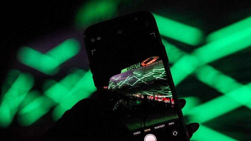 Eine Hand hält ein Smartphone und filmt oder fotografiert eine beleuchtete Arena. Auf dem Display des Smartphones ist die Aufschrift "VW ARENA" zu erkennen. Der Hintergrund zeigt grüne Lichtstreifen, die ein abstraktes Muster bilden.