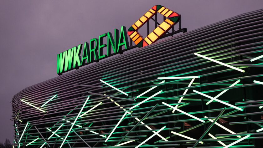 VWK Arena, ein Fußballstadion, bei Nacht. Das Gebäude ist mit grünen und weißen Lichtern beleuchtet, die ein Netzwerkmuster auf der Fassade erzeugen. Das Schild 'VWK ARENA' ist in grünen Buchstaben mit einem gelb-rot-grünen Logo daneben prominent auf dem Dach angebracht.