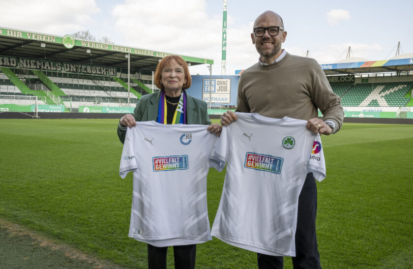 Zwei Personen stehen auf einem Fußballfeld und halten jeweils ein weißes Trikot mit dem Aufdruck "#VIELFALT GEWINNT" in die Kamera. Im Hintergrund sind die grünen Tribünen des Stadions mit Werbebannern zu sehen. Die eine Person ist eine ältere Frau mit roter Kurzhaarfrisur, die einen grünen Blazer und eine bunte Schal trägt. Die andere Person ist ein Mann mit Glatze und Brille, der ein graues Langarmshirt trägt. Die Szene findet im Rahmen einer Medientermin-Kampagne zur Präsentation eines Sondertrikots statt.