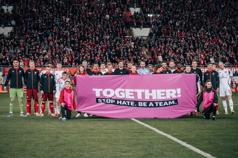 Zwei Fußballmannschaften stehen auf dem Spielfeld und halten ein großes pinkes Banner mit der Aufschrift "TOGETHER! STOP HATE. BE A TEAM." Die Spieler und Spielerinnen sind in ihren Teamuniformen gekleidet und posieren für ein Gruppenfoto vor einer großen Zuschauermenge im Stadion.