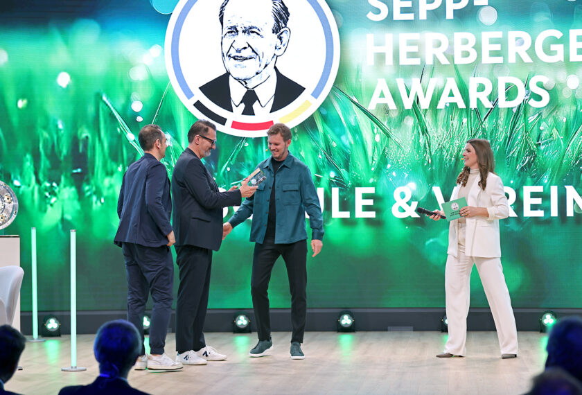 Auf der Bühne der Sepp-Herberger-Awards stehen vier Personen, darunter ein Preisträger, der einen Award erhält. Im Hintergrund ist ein großes Logo mit dem Porträt von Sepp Herberger zu sehen. Die Veranstaltung scheint sich auf die Kategorien "Klub & Verein" zu konzentrieren.