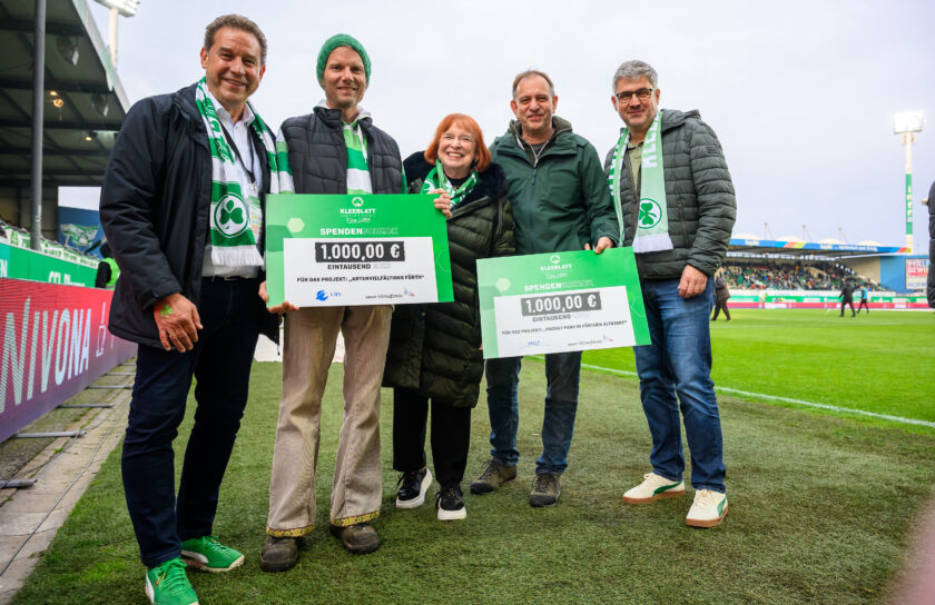 Gruppe von Personen bei einer symbolischen Scheckübergabe auf einem Fußballfeld. Sie halten zwei Schecks über jeweils 1.000,00 Euro für Umweltprojekte in Fürth. Die Personen tragen grüne Schals und Kleidung, was auf die Unterstützung des Fußballvereins SpVgg Greuther Fürth hinweist. Im Hintergrund sind Tribünen und Werbebanner zu sehen.