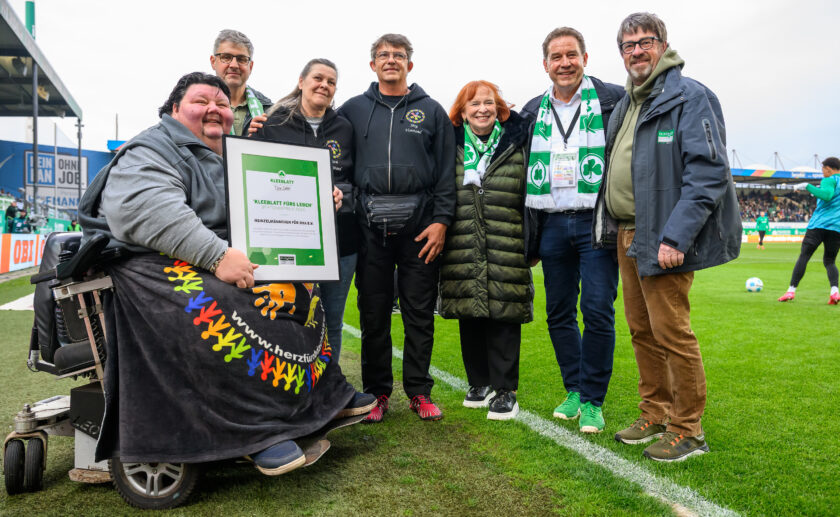 Gruppe von Menschen auf einem Fußballfeld, darunter eine Person im Rollstuhl, die eine Urkunde hält. Die Urkunde trägt den Titel "Kleeblatt fürs Leben" und ist an die Heinzelmännchen für OHA e.V. vergeben. Die Gruppe besteht aus Markus Braun, dem Bürgermeister der Stadt Fürth, Ingrid Hofmann, der geschäftsführenden Alleingesellschafterin der I.K. Hofmann GmbH, und weiteren Personen. Alle lächeln und tragen Kleidung mit grün-weißen Farben, was auf die Verbindung zum Fußballverein SpVgg Greuther Fürth hinweist. Im Hintergrund sind Teile des Stadions und einige Zuschauer zu sehen.