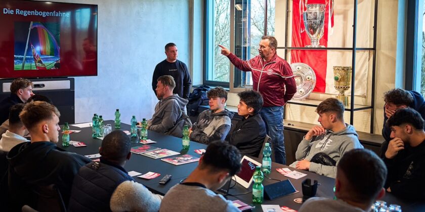 Eine Gruppe von Personen nimmt an einem Workshop teil, der von einem Referenten geleitet wird. Der Referent steht und zeigt auf eine Präsentation auf einem großen Bildschirm, auf dem die Regenbogenfahne zu sehen ist. Die Teilnehmer sitzen an Tischen und hören aufmerksam zu. Im Hintergrund sind verschiedene Trophäen und Banner zu sehen, darunter auch solche des FC Bayern. Auf den Tischen stehen Flaschen mit Wasser und Informationsmaterialien.