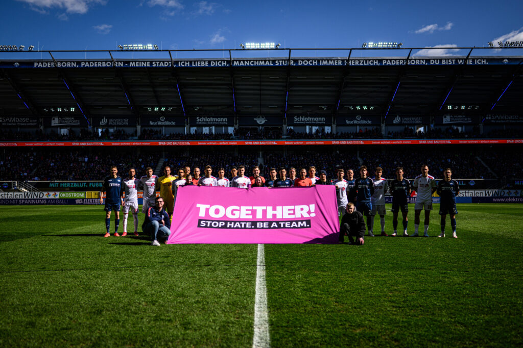 Zwei Fußballmannschaften stehen auf dem Rasen eines Stadions und halten gemeinsam ein pinkes Banner mit der Aufschrift 'TOGETHER! STOP HATE. BE A TEAM.' Die Spieler sind in verschiedenen Trikots gekleidet, und im Hintergrund sind die Tribünen mit Zuschauern zu sehen. Das Bild wurde während eines Zweitliga-Spiels zwischen SC Paderborn 07 und 1. FC Köln aufgenommen.