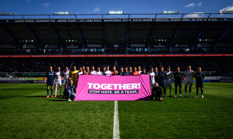Zwei Fußballmannschaften stehen auf dem Rasen eines Stadions und halten gemeinsam ein pinkes Banner mit der Aufschrift 'TOGETHER! STOP HATE. BE A TEAM.' Die Spieler sind in verschiedenen Trikots gekleidet, und im Hintergrund sind die Tribünen mit Zuschauern zu sehen. Das Bild wurde während eines Zweitliga-Spiels zwischen SC Paderborn 07 und 1. FC Köln aufgenommen.