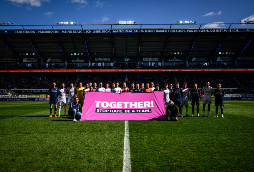 Zwei Fußballmannschaften stehen auf dem Rasen eines Stadions und halten gemeinsam ein pinkes Banner mit der Aufschrift 'TOGETHER! STOP HATE. BE A TEAM.' Die Spieler sind in verschiedenen Trikots gekleidet, und im Hintergrund sind die Tribünen mit Zuschauern zu sehen. Das Bild wurde während eines Zweitliga-Spiels zwischen SC Paderborn 07 und 1. FC Köln aufgenommen.