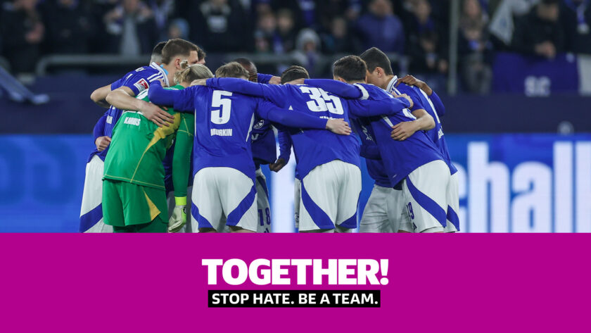 Eine Gruppe von Fußballspielern in blauen Trikots und einem Torwart in grünem Trikot umarmen sich auf dem Spielfeld. Im Vordergrund steht der Text "TOGETHER! STOP HATE. BE A TEAM." auf pinkem Hintergrund.