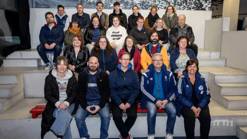 Eine Gruppe von Menschen sitzt auf den Stufen eines Stadions und lächelt in die Kamera. Sie tragen verschiedene Jacken und Kleidung, darunter einige mit Logos und Aufschriften. Im Hintergrund ist eine große Schwarz-Weiß-Fotografie von Fußballfans zu sehen. Die Szene findet im Parkstadion statt, wie auf den Stufen zu lesen ist.