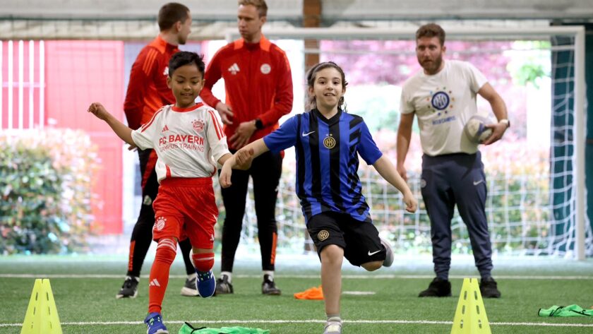 Zwei Kinder im Fußballtraining, eines im Trikot des FC Bayern München und das andere im Trikot von Inter Mailand, führen eine Übung mit Hütchen auf dem Platz aus. Im Hintergrund stehen Trainer in roten und weißen Outfits sowie ein Trainer in einem weißen T-Shirt mit dem Logo des WSV.