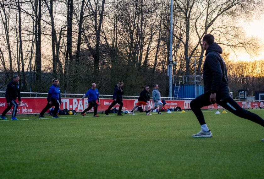 Eine Gruppe von Menschen absolviert ein Fußballtraining auf einem Rasenplatz bei Sonnenuntergang. Die Personen sind in Sportkleidung gekleidet und führen verschiedene Übungen aus. Im Hintergrund sind Bäume und eine Werbebande mit der Aufschrift "Besser offen, besser leben" zu sehen.