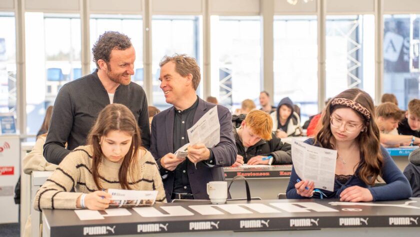 Gruppe von Menschen bei einer Veranstaltung in einer Arena. Zwei Männer und zwei Frauen sitzen an einem Tisch mit Puma-Logo und füllen Formulare aus. Im Hintergrund sind weitere Personen zu sehen, die ebenfalls beschäftigt sind. Die Atmosphäre wirkt geschäftig und konzentriert.