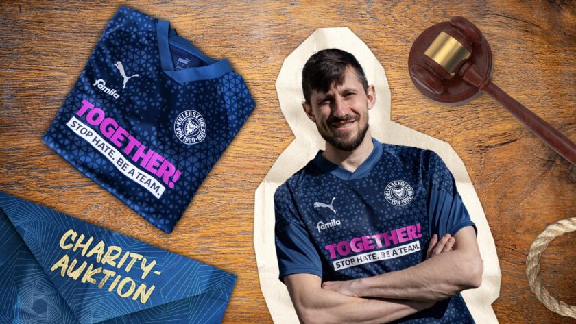 Ein lächelnder Mann in einem blauen Trikot mit der Aufschrift "TOGETHER! STOP HATE. BE A TEAM." sitzt auf einem Richterstuhl. Neben ihm liegt ein Trikot mit ähnlichem Design und ein Holzhammer, der an eine Auktion erinnert. Der Hintergrund ist ein Holztisch mit einem Schild, auf dem "CHARITY-AUKTION" steht.