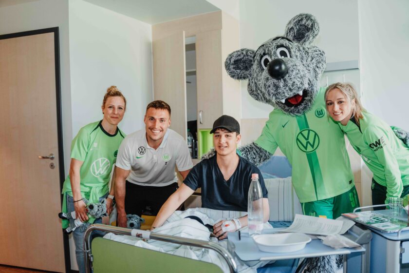 Gruppe von Menschen und einer Maske in einem Krankenhauszimmer. Eine Person liegt im Bett, während andere stehen und lächeln. Alle tragen grüne und weiße Kleidung mit Volkswagen-Logos. Eine Person hält ein Stofftier. Im Hintergrund ist ein Türrahmen und ein Tisch mit einer Wasserflasche und Papieren zu sehen.