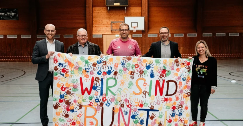 Fünf Personen stehen in einer Sporthalle und halten gemeinsam ein buntes Transparent. Das Transparent zeigt viele verschiedene Handabdrücke in verschiedenen Farben und die Aufschrift "WIR SIND BUNT". Die Personen lächeln und wirken zufrieden. Die Halle hat Holzwände und einen Basketballkorb im Hintergrund.