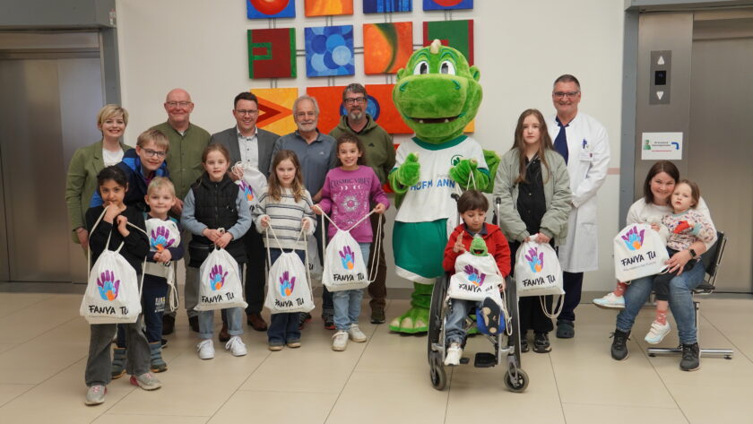 Eine Gruppe von Kindern und Erwachsenen steht in einem Flur vor einer bunten Wand. Sie halten weiße Stoffbeutel mit dem Logo "FANYA TU Foundation for Future". In der Mitte der Gruppe steht eine Person im Kostüm eines grünen Dinosauriers und trägt ein T-Shirt mit der Aufschrift "Hofmann Ann". Die Gruppe scheint für ein Gruppenfoto zu posieren.
