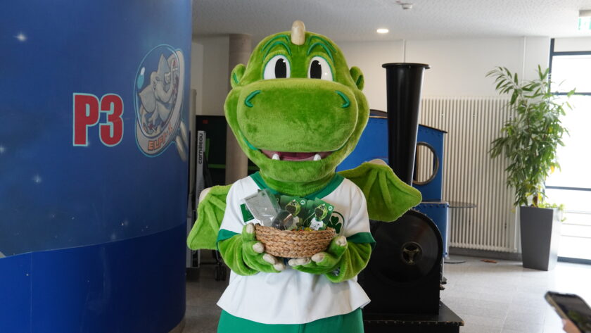 Ein grüner Maskottchen-Dino in einem weißen Trikot mit grünen Akzenten steht vor einer blauen Wand mit der Aufschrift "P3" und einem Logo. Das Maskottchen hält einen Korb mit verschiedenen Gegenständen, darunter eine Flasche und ein Spielzeug-Dino. Im Hintergrund sind ein schwarzer Mülleimer und eine Pflanze in einem Topf zu sehen.