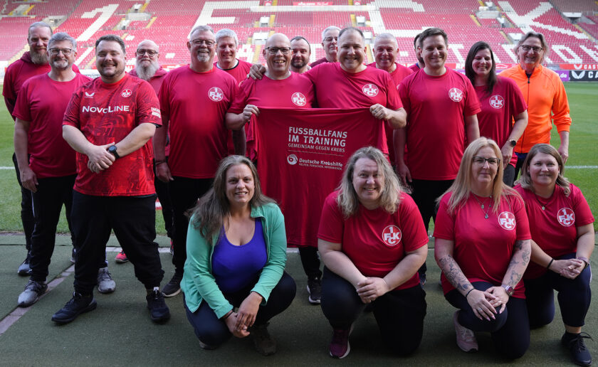 Eine Gruppe von Menschen in roten T-Shirts mit dem Logo des 1. FC Köln steht und kniet auf einem Fußballfeld. Sie halten ein rotes Tuch mit der Aufschrift "FUSSBALLFANS IM TRAINING GMEINSAM GEGEN KREBS" und dem Logo der Deutschen Krebshilfe. Im Hintergrund sind die roten Sitzplätze des Stadions zu sehen.