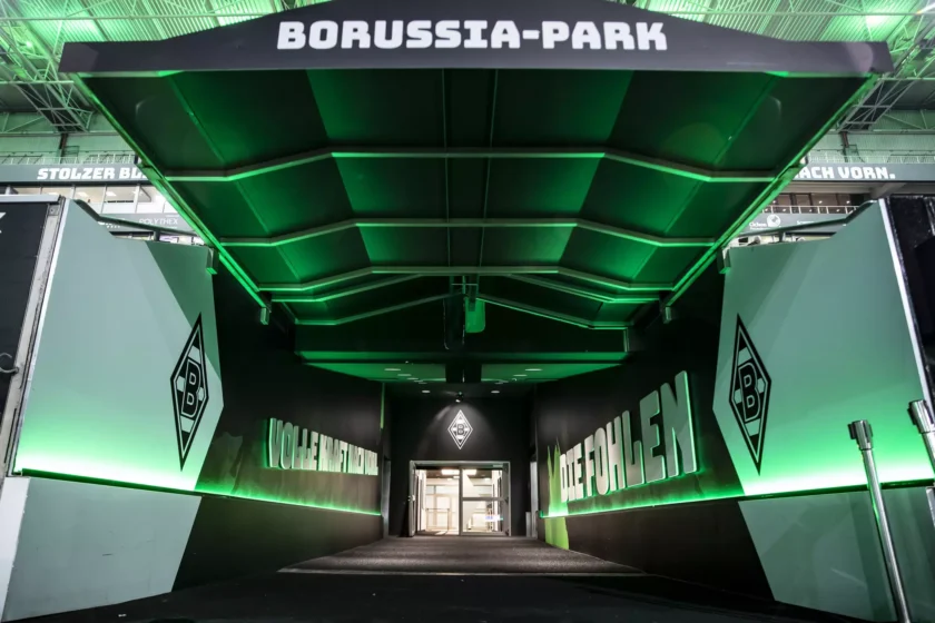 Eingangstunnel des Borussia-Parks, Heimatstadion von Borussia Mönchengladbach, mit grün beleuchteten Wänden und dem Schriftzug 'VfL WOHLEN' sowie dem Logo des Vereins.
