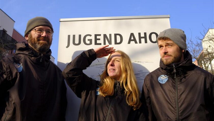 Drei Personen stehen vor einem Schild mit der Aufschrift "JUGEND AHOI". Sie tragen warme Kleidung und lächeln in die Kamera. Die Person in der Mitte hält ihre Hand an die Stirn, als ob sie in die Ferne schaut. Die Szene wirkt fröhlich und entspannt.