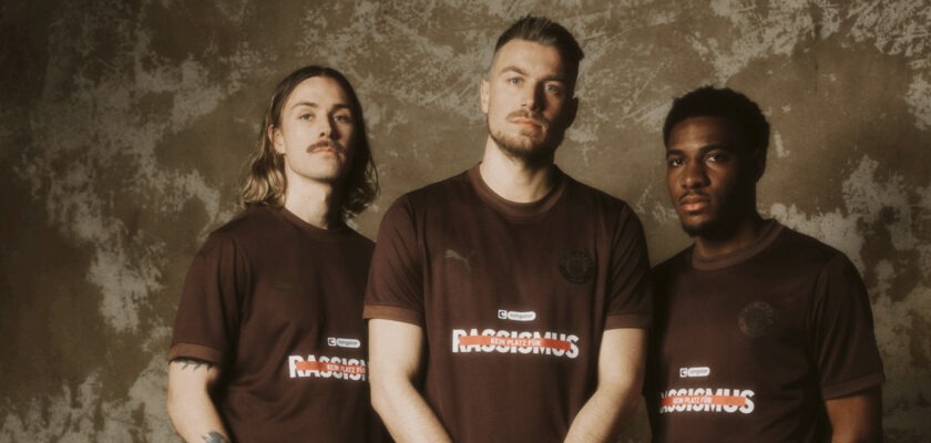 Drei Personen stehen nebeneinander vor einer grauen Wand und tragen braune T-Shirts mit dem Aufdruck "Rassismus". Die T-Shirts haben auch Logos und andere Inschriften, die auf eine Kampagne gegen Rassismus hinweisen. Die Personen haben unterschiedliche Hautfarben und Bärte, was die Vielfalt und Einheitlichkeit der Botschaft unterstreicht.