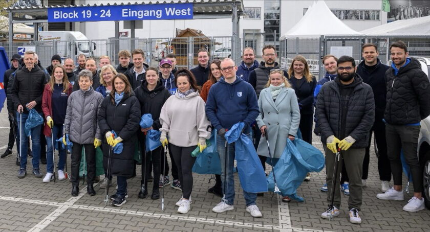 Gruppe von Menschen beim freiwilligen Müllsammeln am Eingang West eines Stadions. Die Teilnehmer tragen warme Kleidung und halten Müllsäcke und Greifzangen in den Händen. Sie stehen vor einem Schild, das "Block 19 - 24 Eingang West" anzeigt.