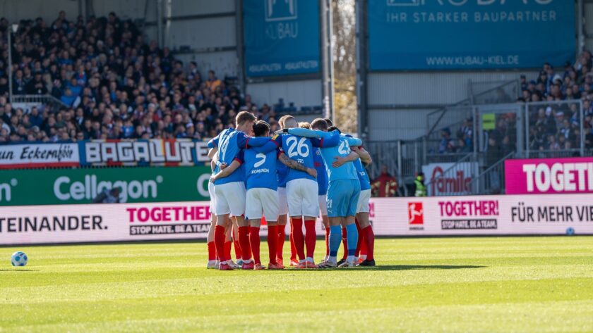 Eine Gruppe von Fußballspielern in blauen Trikots und roten Stutzen steht im Mannschaftskreis auf dem Rasen. Sie haben sich im Kreis zusammengeschlossen und umarmen sich. Im Hintergrund sind Zuschauer auf den Tribünen zu sehen, sowie Werbebanner mit der Aufschrift "TOGETHER - FÜR MEHR VIELFALT. STOP HATE. BEAT RACISM.