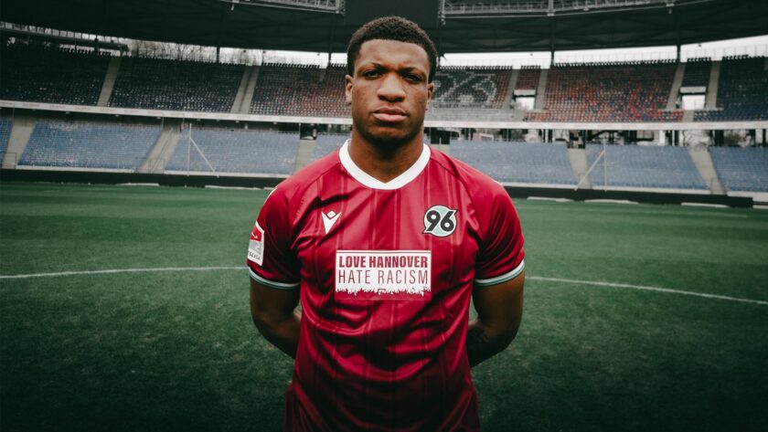 Ein Fußballspieler steht auf einem Fußballfeld in einem Stadion. Er trägt ein rotes Trikot mit dem Logo von Hannover 96 und dem Aufdruck "LOVE HANNOVER HATE RACISM". Im Hintergrund sind die leeren Tribünen des Stadions zu sehen.