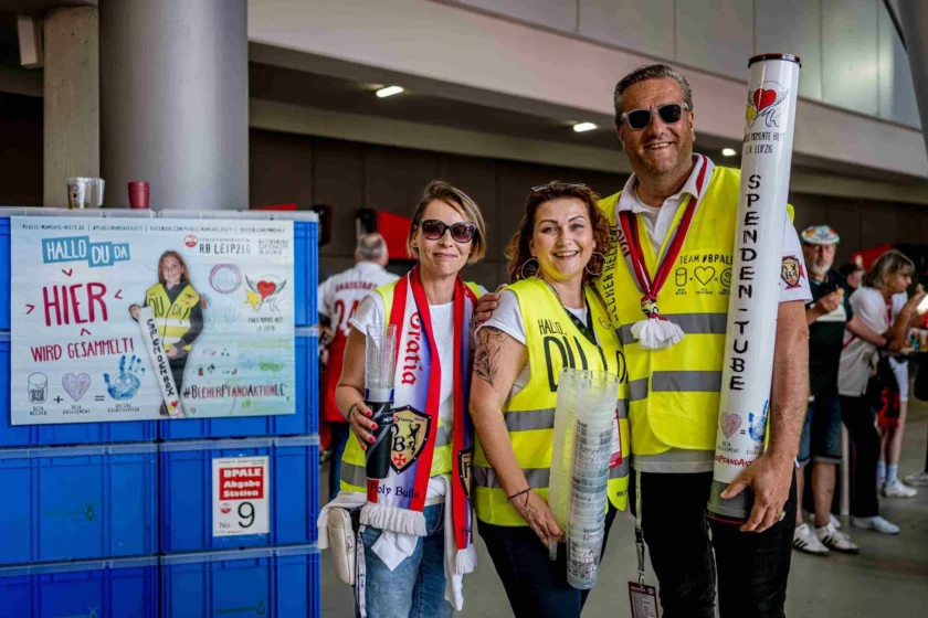 Drei Personen in gelben Westen stehen vor einer Sammelstation für Pfandbecher in der Red Bull Arena. Sie halten leere Pfandbecher in den Händen und lächeln in die Kamera. Im Hintergrund ist ein Schild mit der Aufschrift "Hallo Du da, hier wird gesammelt!" zu sehen. Die Aktion findet im Rahmen der "BecherPfandAktionLC" statt.