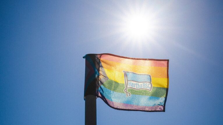 Ein Regenbogen-Flagge mit dem Logo des Hertha BSC weht im Wind an einem klaren, sonnigen Himmel.